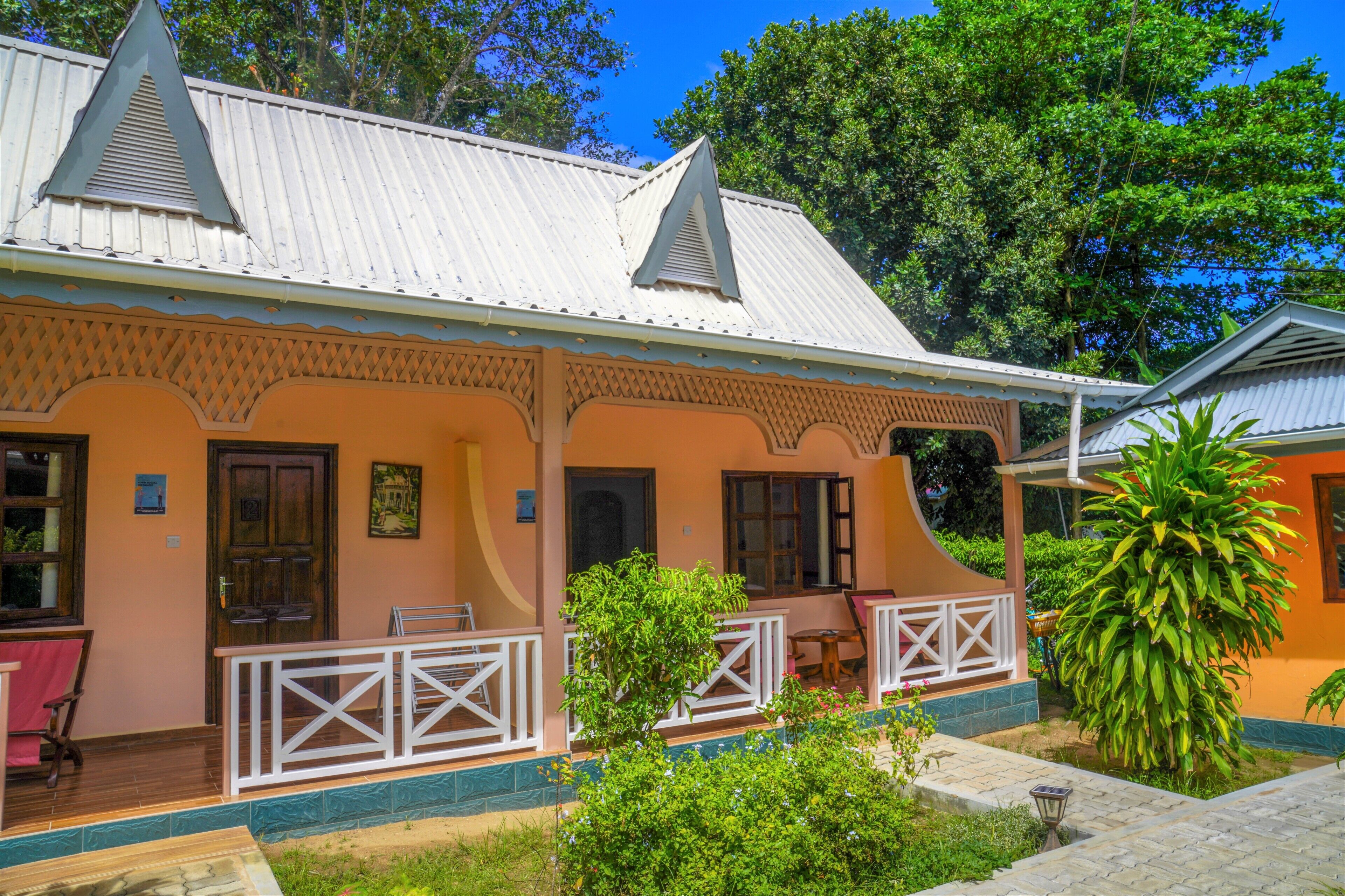 A two-story building with a porch, a wooden door, and a small garden area with a palm tree.