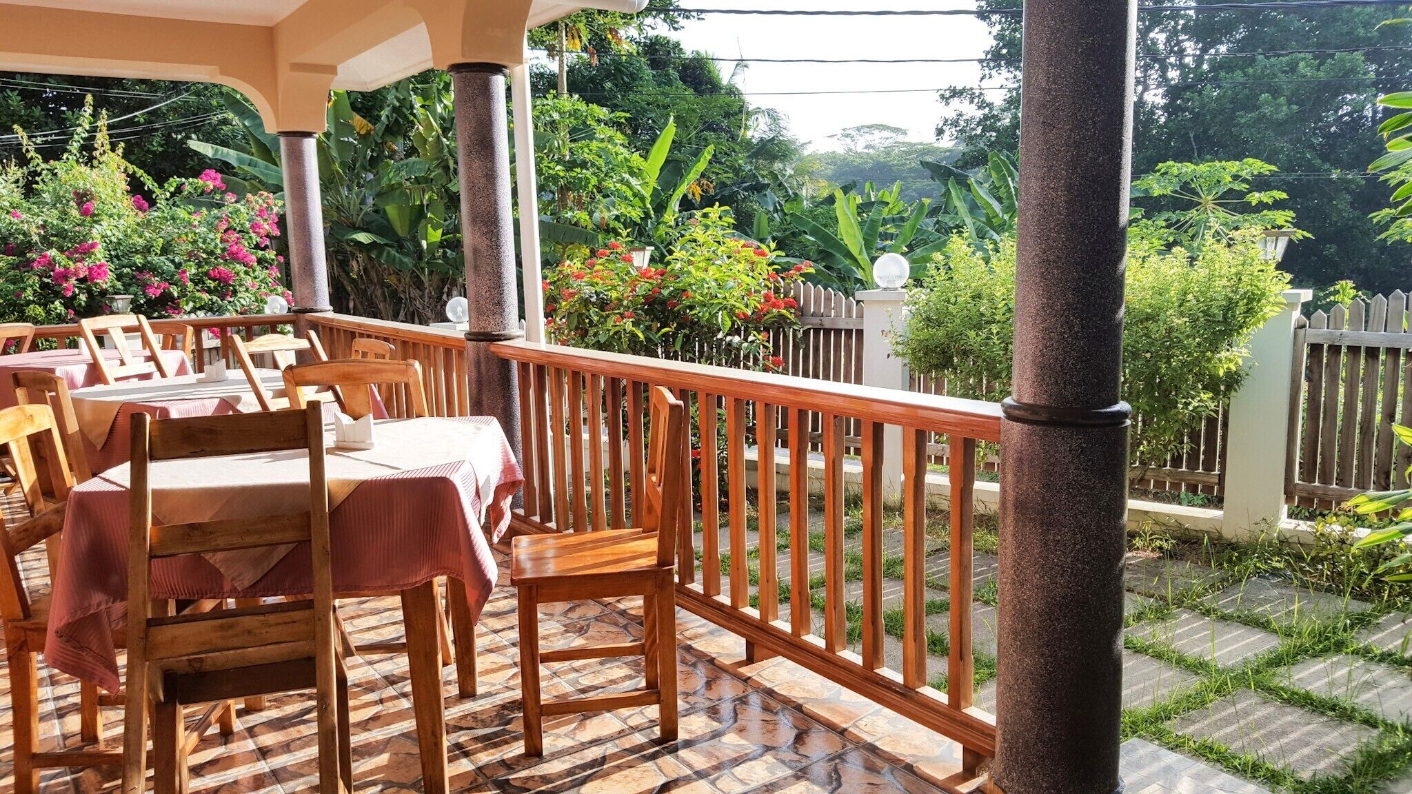 An outdoor patio with wooden tables and chairs, surrounded by a wooden fence and lush greenery.