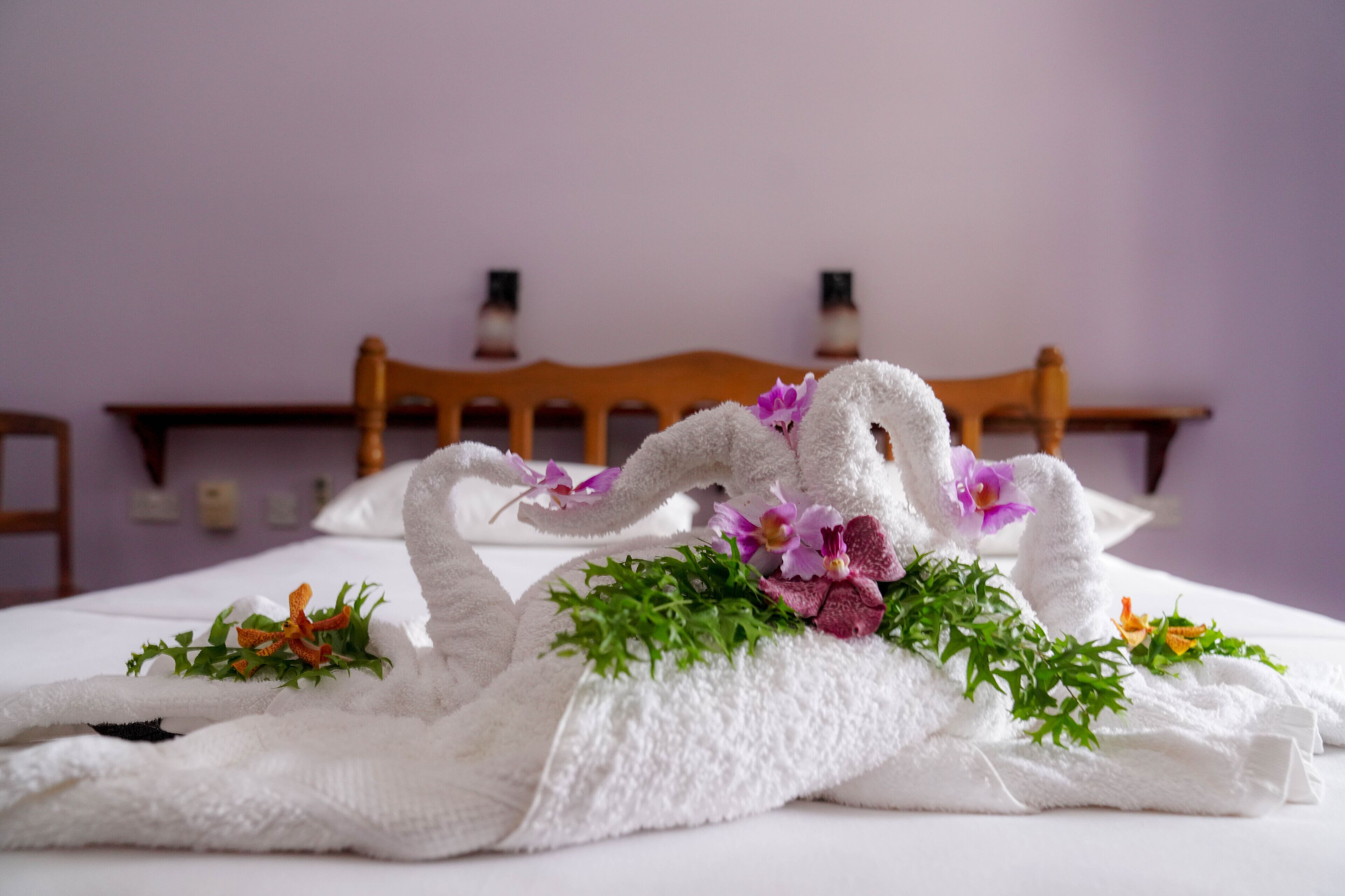 Una cama cuidadosamente arreglada con una toalla blanca dispuesta en forma de cisne, decorada con flores moradas y follaje verde.