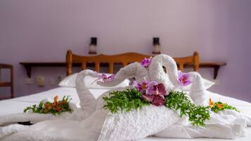Una cama cuidadosamente arreglada con una toalla blanca dispuesta en forma de cisne, decorada con flores moradas y follaje verde.