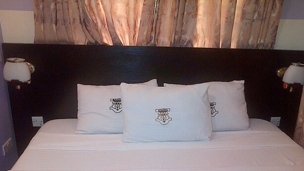 A bed with white bedding and three pillows featuring a crest logo. - Esado Suites and Conference Oregun (Lagos)