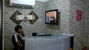 A reception area with a stone wall, a flat-screen TV mounted on the wall, and a receptionist standing behind a glass desk. - Esado Suites and Conference Oregun (Lagos)