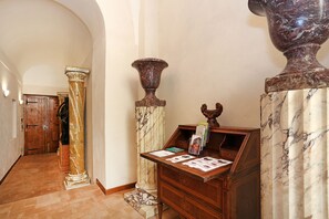 Un pasillo con columnas de mármol, un escritorio de madera con folletos y una puerta de madera al fondo.