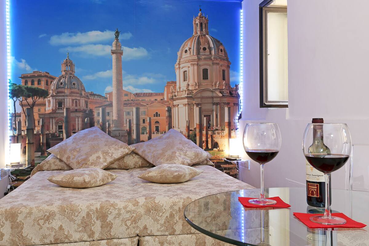 Ein Bett mit gemustertem Überwurf und Kissen, ein Glastisch mit zwei Weingläsern und einer Flasche, sowie eine Wandmalerei eines historischen Gebäudes im Hintergrund.