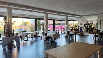 A spacious indoor area with multiple tables and chairs, large windows with a view of the outside, and a variety of plants and bottles on a central table.