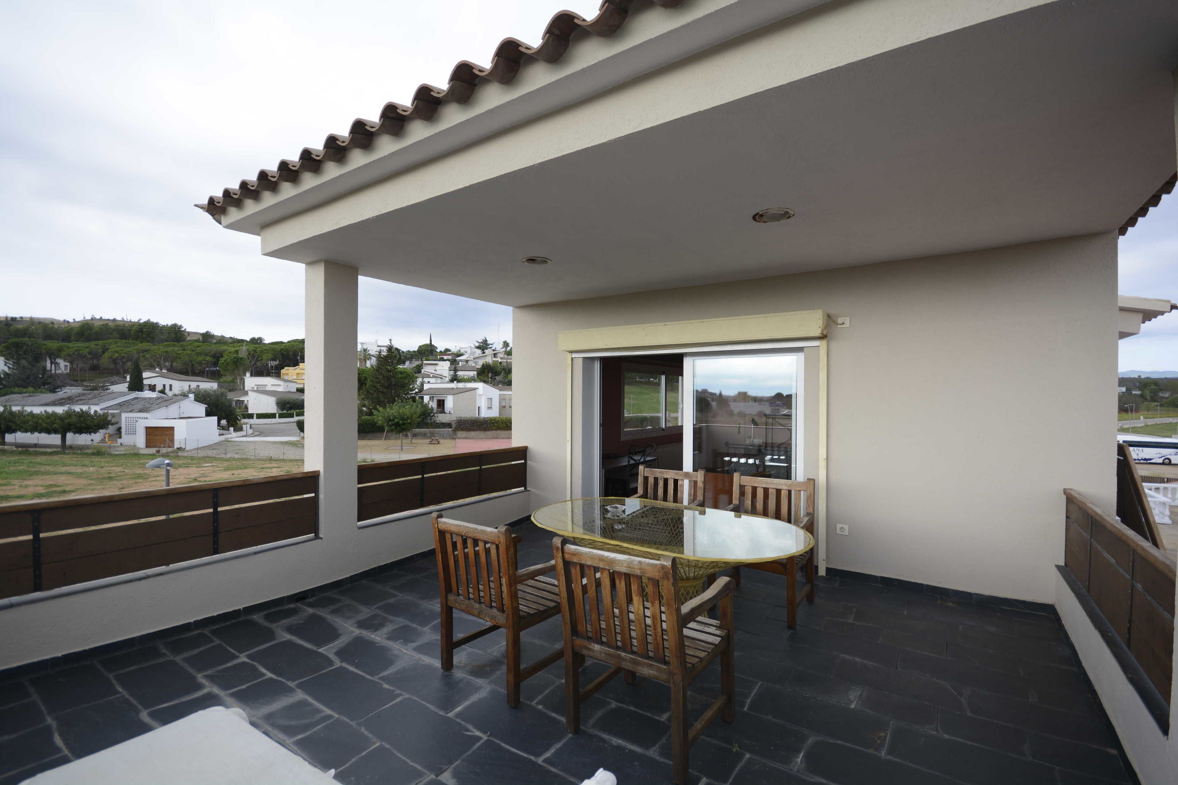 A covered patio with a glass table and chairs, a view of the surrounding neighborhood, and a tiled floor.