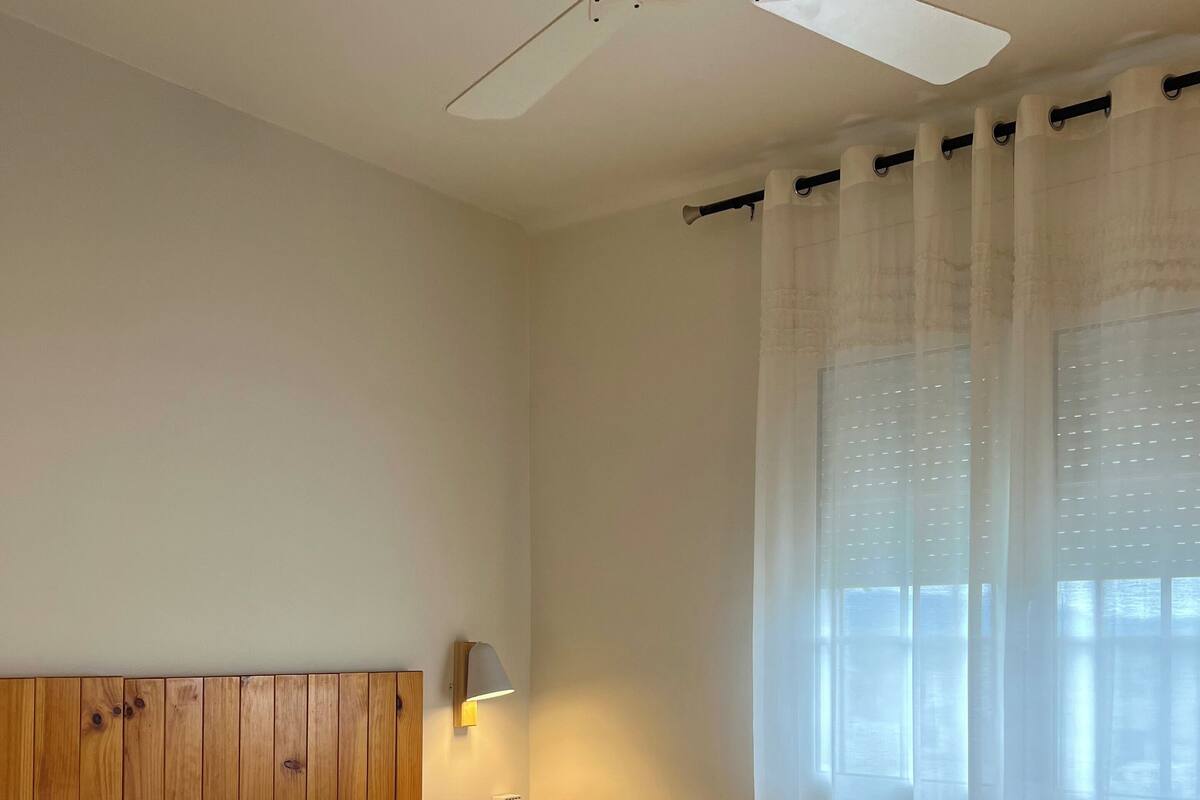 A bedroom with two beds, a wooden headboard, a ceiling fan, a nightstand with a lamp, and sheer curtains.