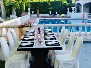 A poolside dining area with a table set for a meal, surrounded by white chairs, and a pool in the background.