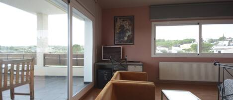 Una sala moderna con un sofá, una mesa de centro de vidrio y un televisor. Hay un ventanal con vistas a edificios y árboles.