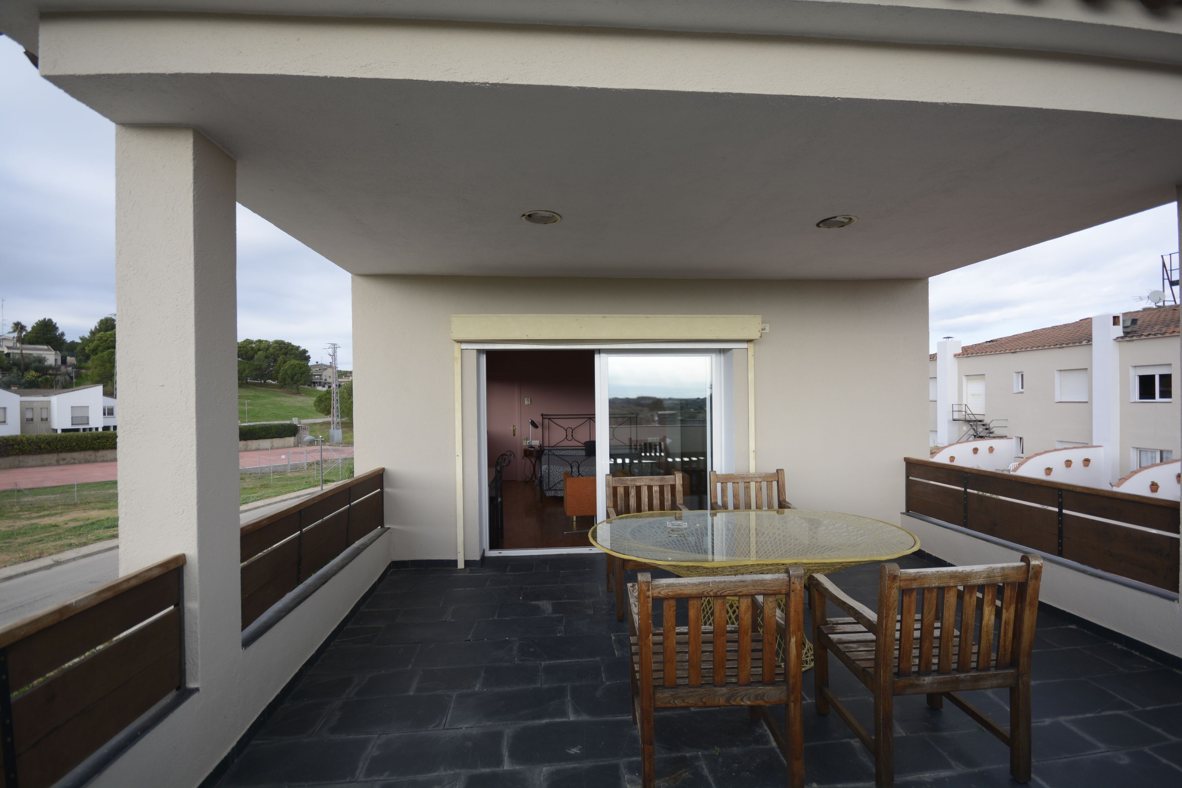 Une terrasse extérieure couverte, avec une table en verre et des chaises en bois, donnant sur la rue, et une porte menant à un espace intérieur.