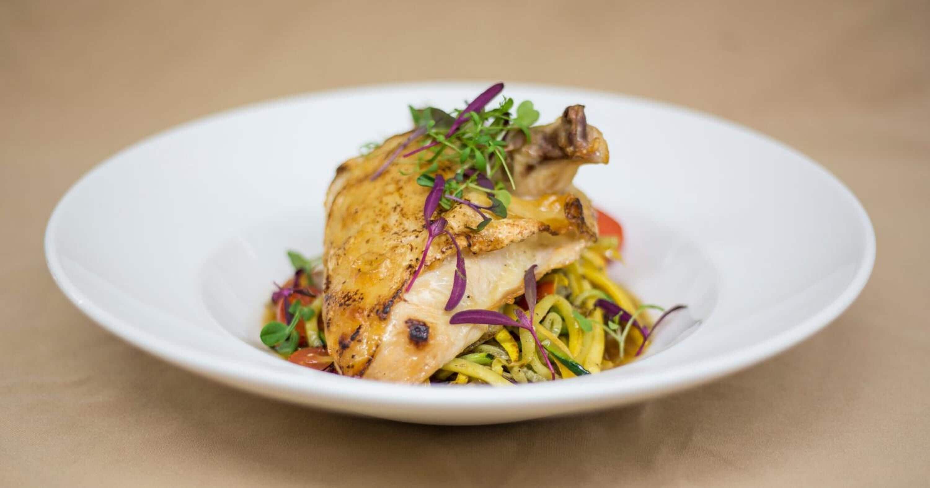 Une assiette blanche contenant un poulet rôti, agrémenté d’herbes violettes et vertes, et accompagné d’un assortiment de légumes.