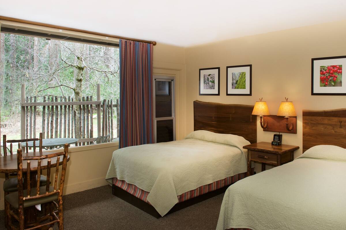 Habitación de hotel con dos camas, cabecero de madera, mesitas de noche con lámparas, ventana con vista a árboles y un comedor con mesa y sillas.