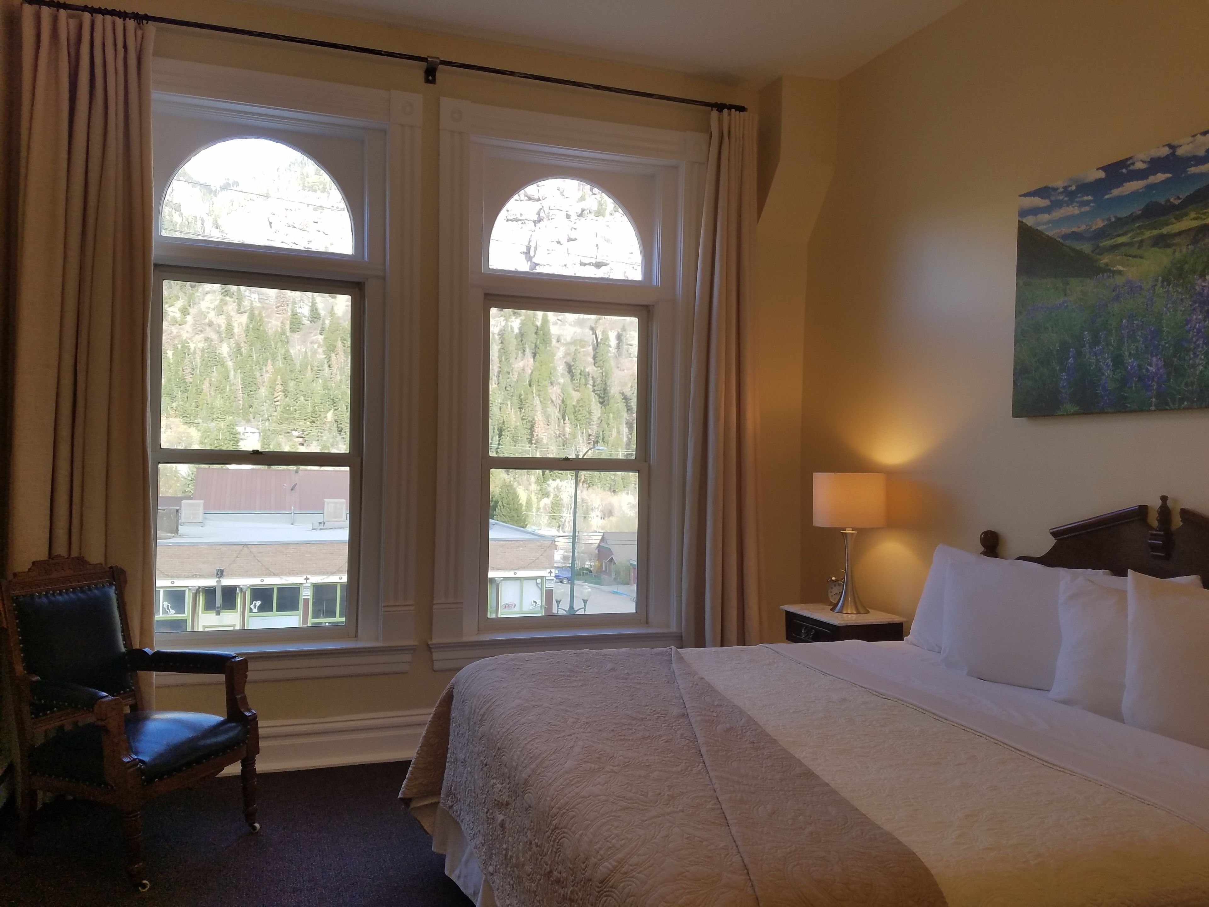 A hotel room with a large bed, two windows with curtains, a chair, a bedside table with a lamp, and a painting on the wall.