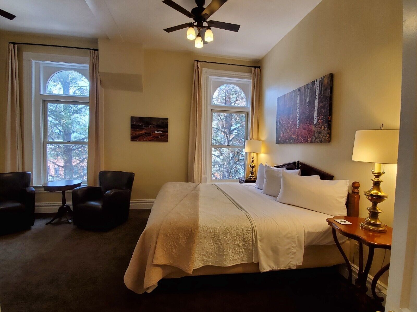 A hotel room with a bed, two chairs, a table, a ceiling fan, two windows, and artwork on the walls.