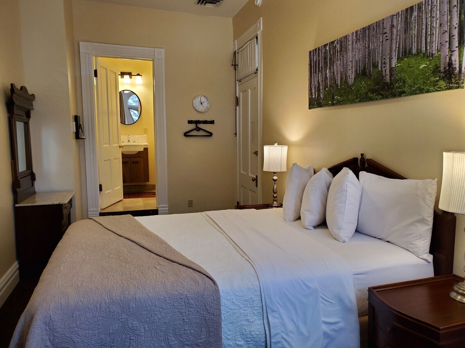 A hotel room with a large bed, a bedside table with a lamp, a wall clock, and a painting of a forest.