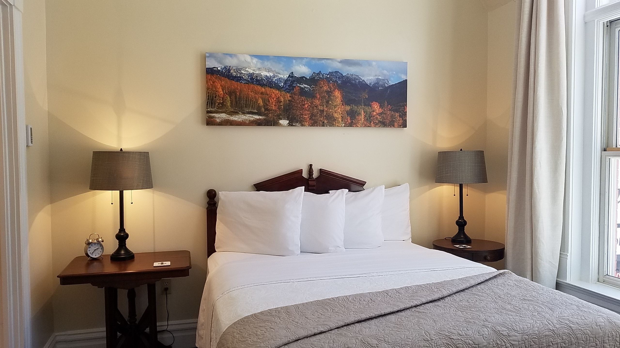 Habitación de hotel con cama, dos mesitas de noche con lámparas, un cuadro de paisaje montañoso y una ventana con cortinas.