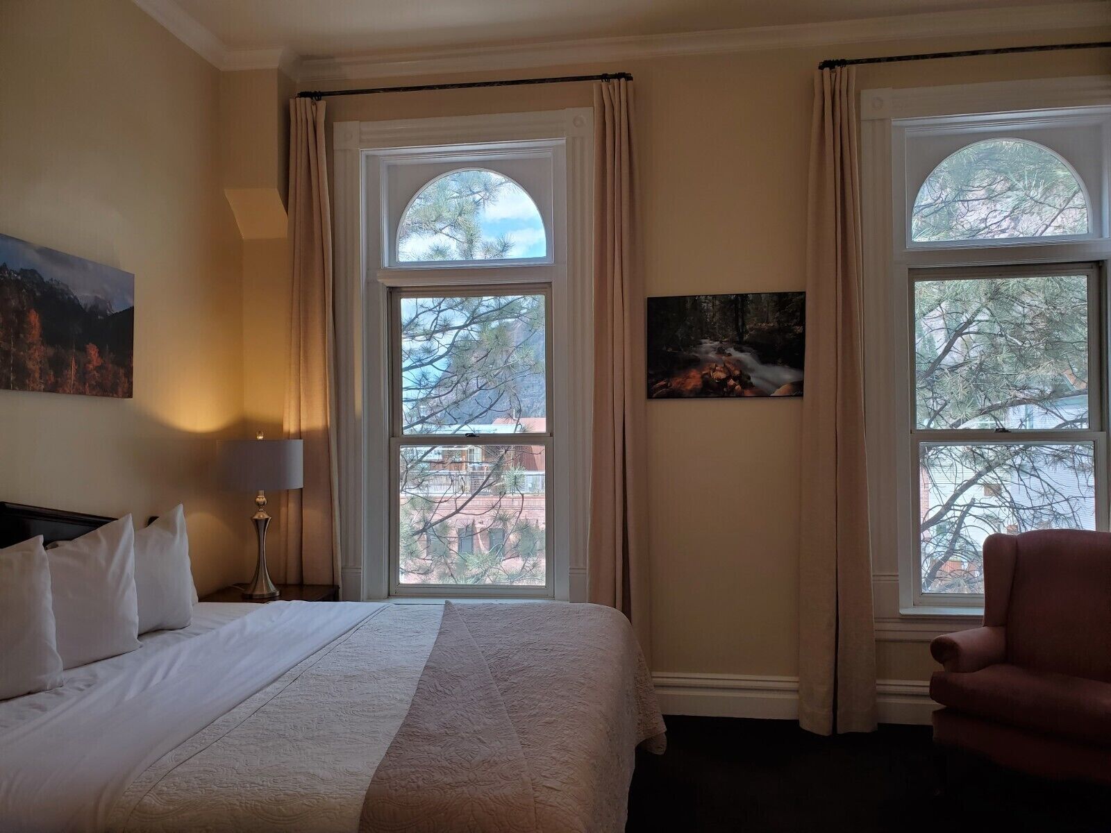 Habitación de hotel con una cama grande, dos ventanas con cortinas, un cuadro en la pared y una silla.