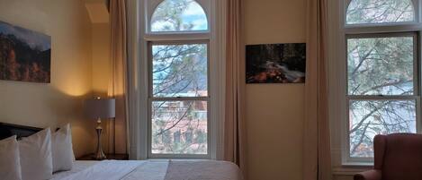 Habitación de hotel con una cama grande, dos ventanas con cortinas, un cuadro en la pared y una silla.