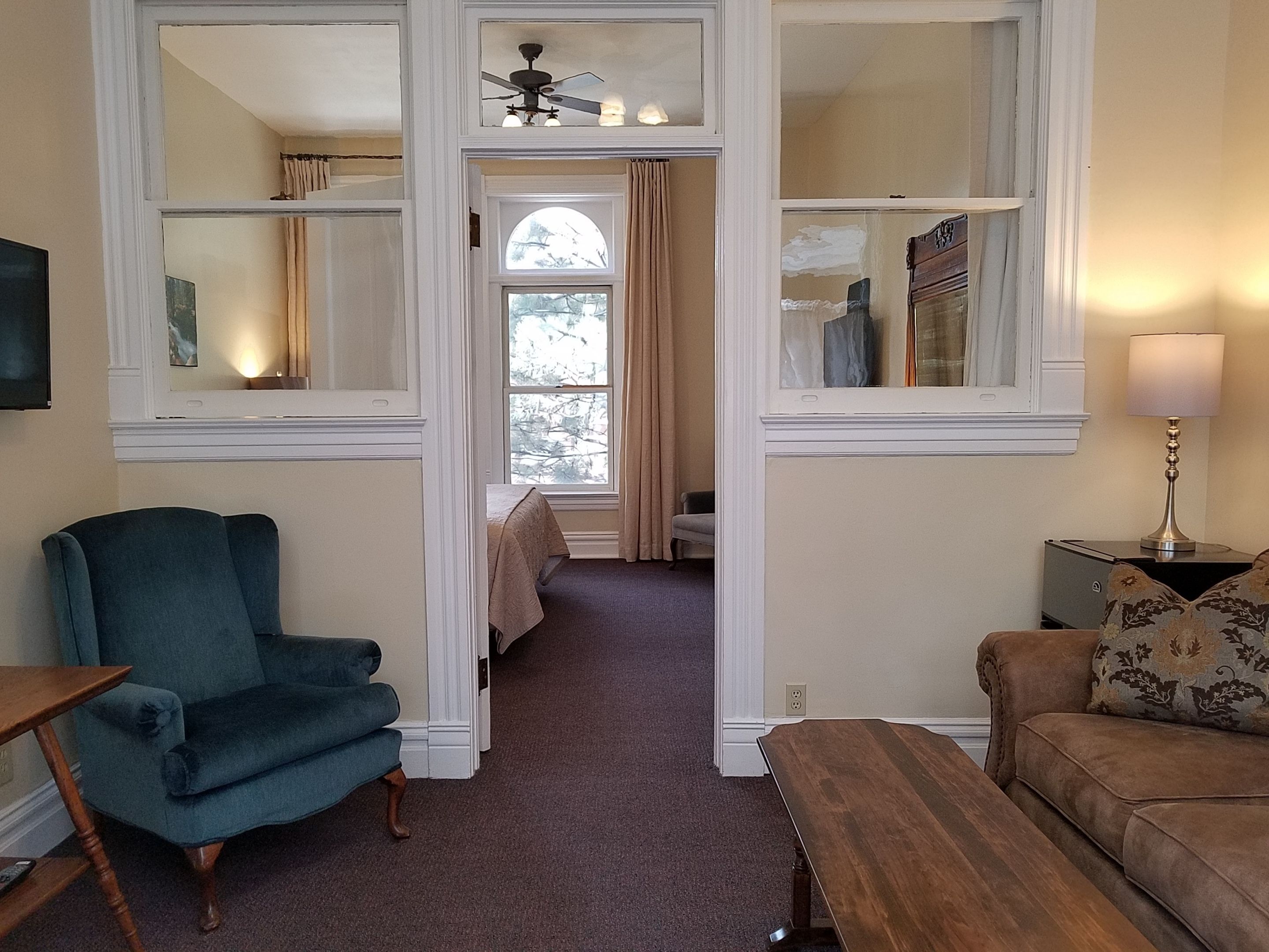 A living room with a blue armchair, a wooden bench, and a brown sofa. There is a ceiling fan and a window with curtains.