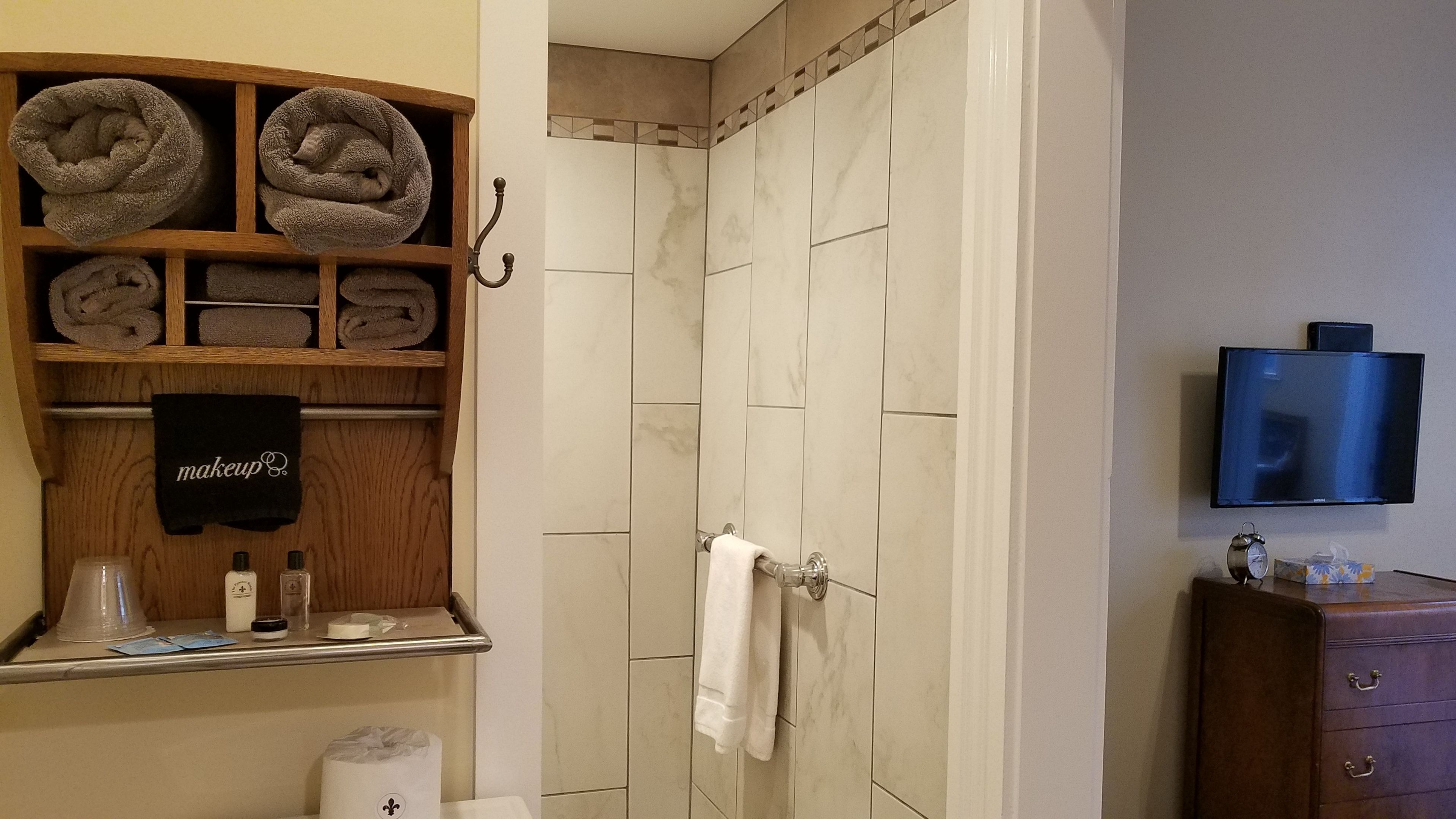 A bathroom with a shower, a wooden shelf with towels, a makeup towel, toiletries, and a TV on the wall.