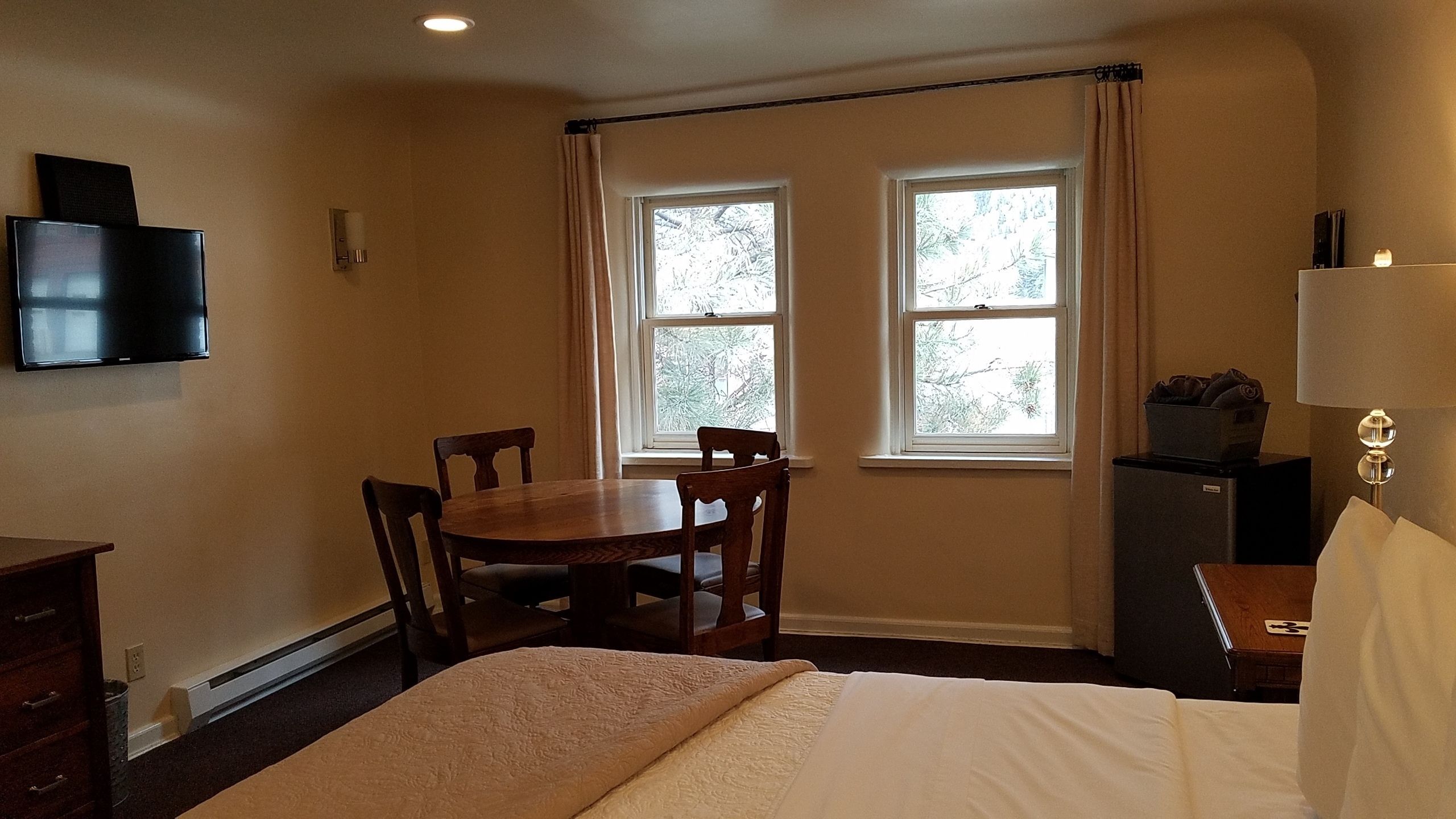 Habitación de hotel con una cama, una mesa y dos sillas, un televisor montado en la pared y dos ventanas con cortinas.