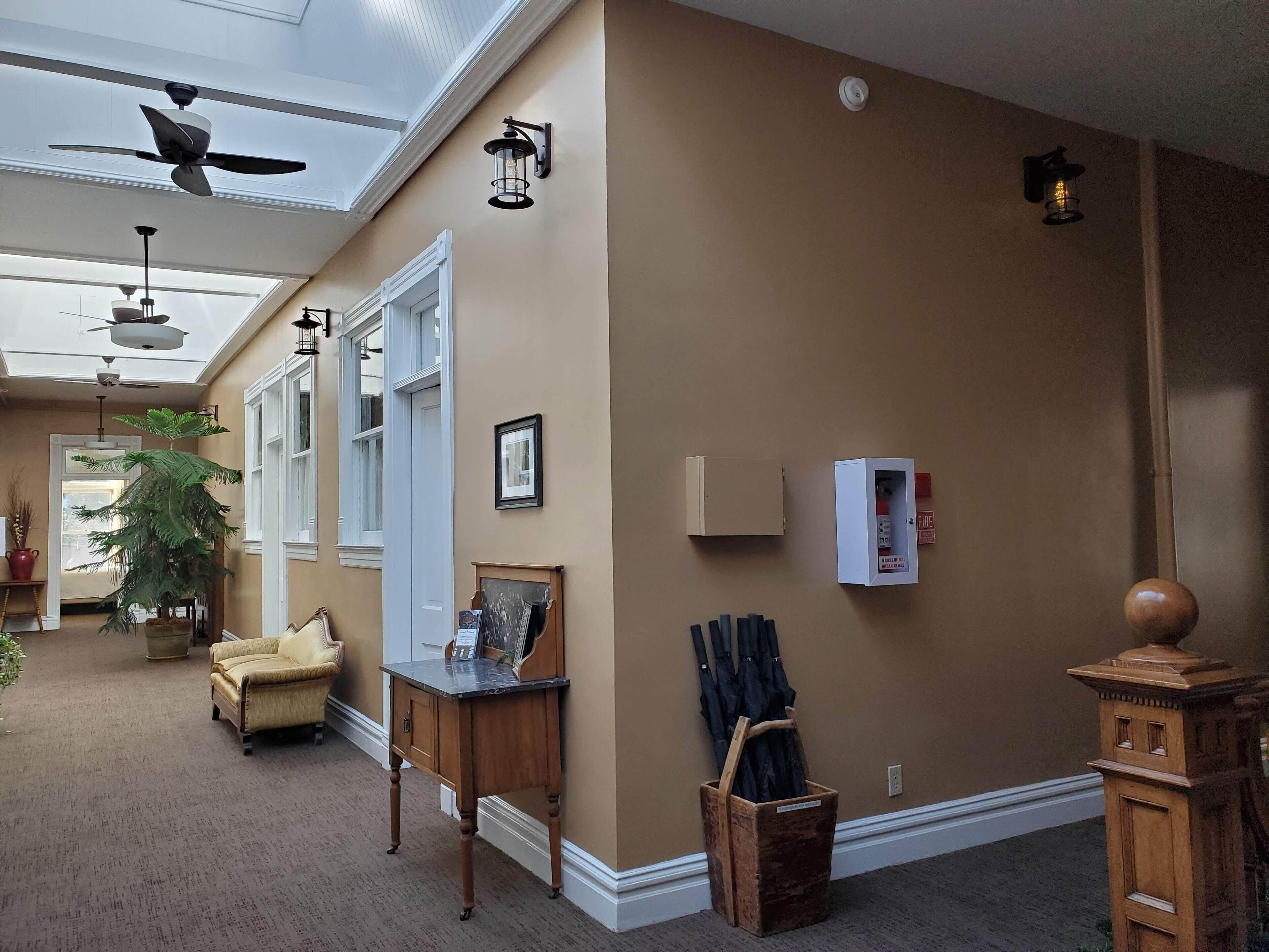 A hallway with a ceiling fan, wall-mounted lights, a fire extinguisher, and a wooden cabinet.