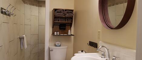 Un baño con inodoro, lavabo y espejo. Hay toallas y artículos de aseo en un estante.