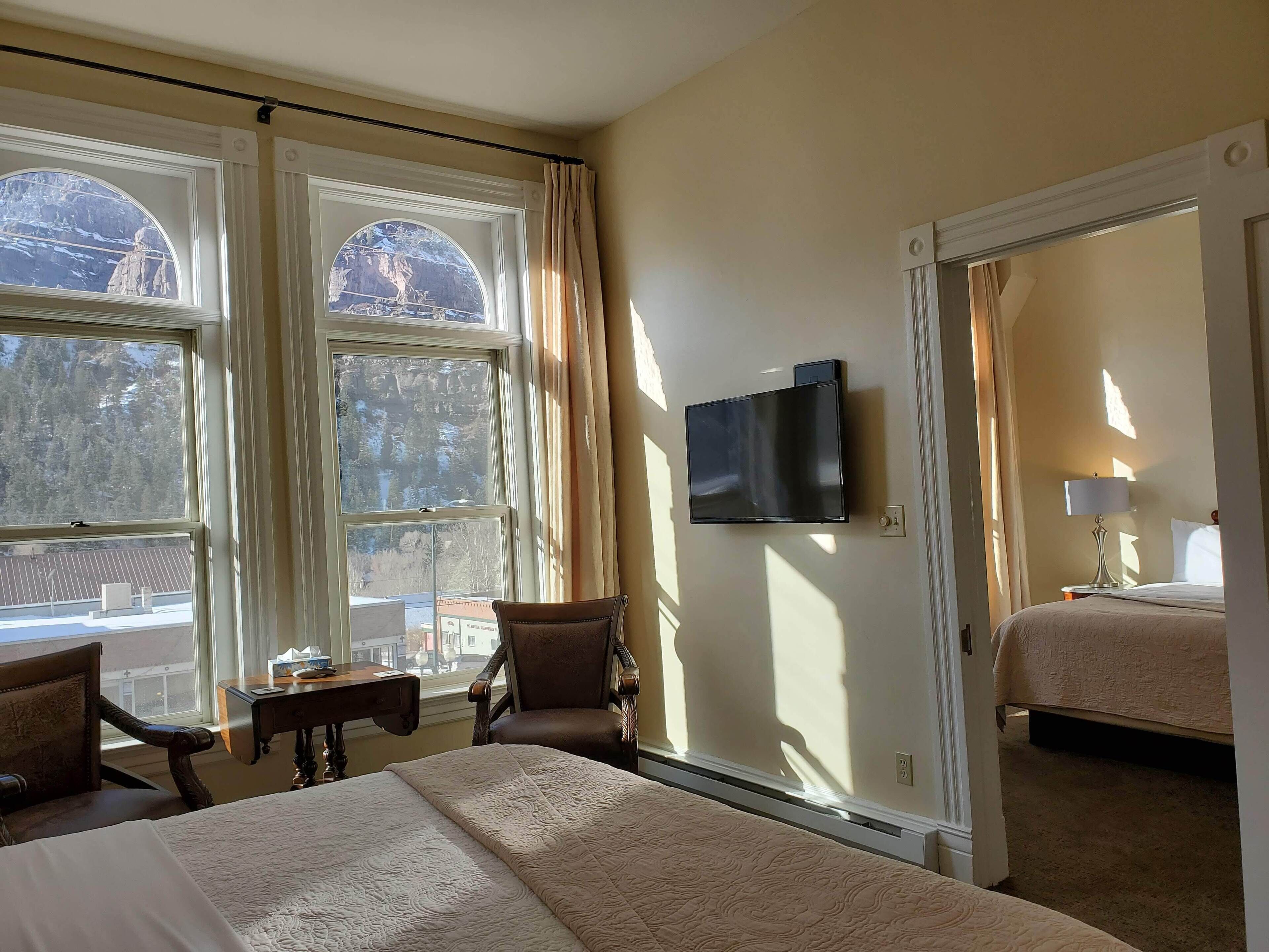 A hotel room with a large bed, a desk, a chair, a TV mounted on the wall, and a view of the outdoors through large windows.