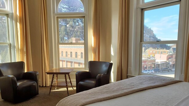 Habitación de hotel con una cama grande, dos sillones, una mesita y amplias ventanas con vistas a un pueblo y montañas.