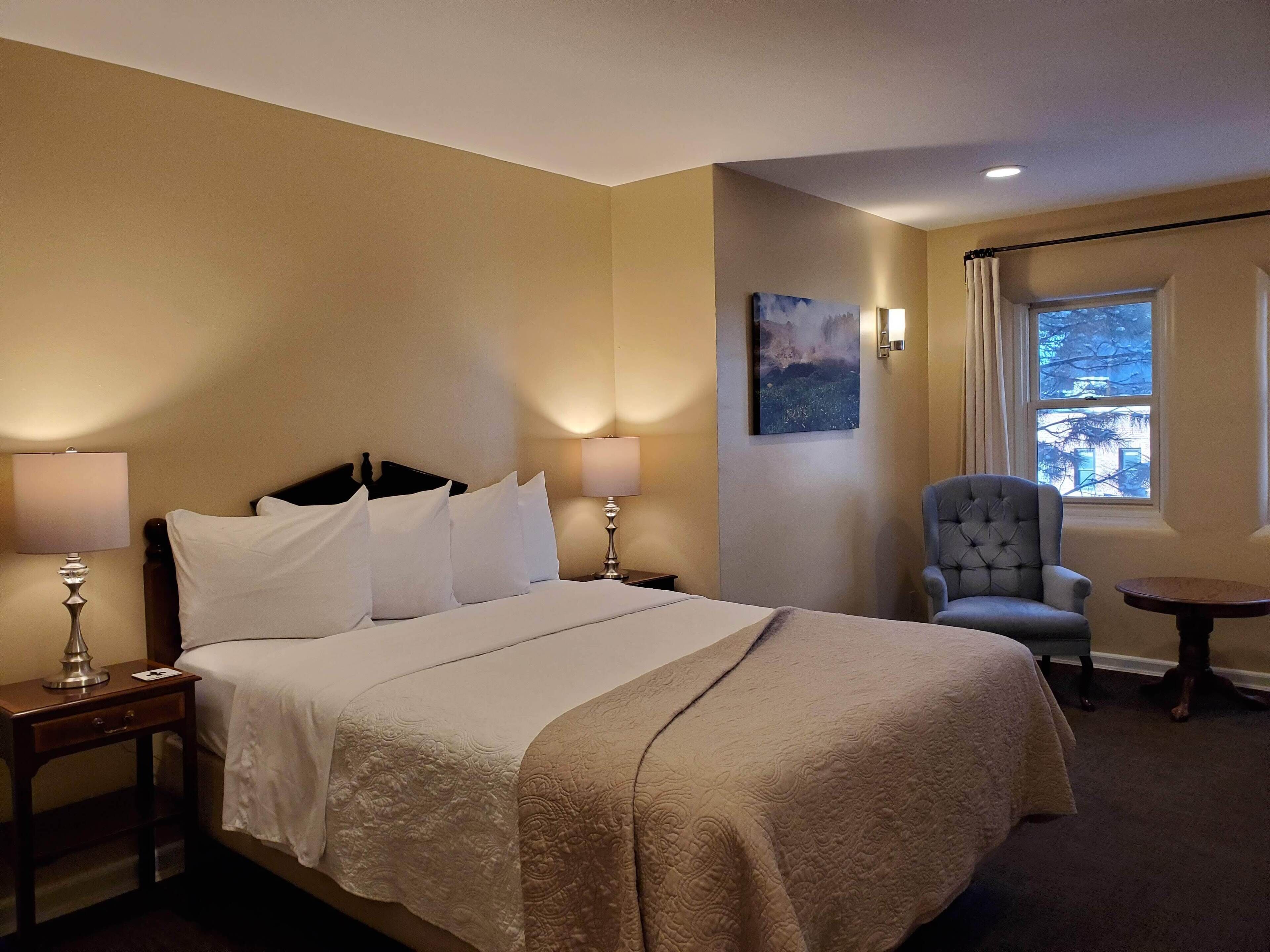 A hotel room with a large bed, two bedside lamps, a chair, a small table, and a window with a view of trees.
