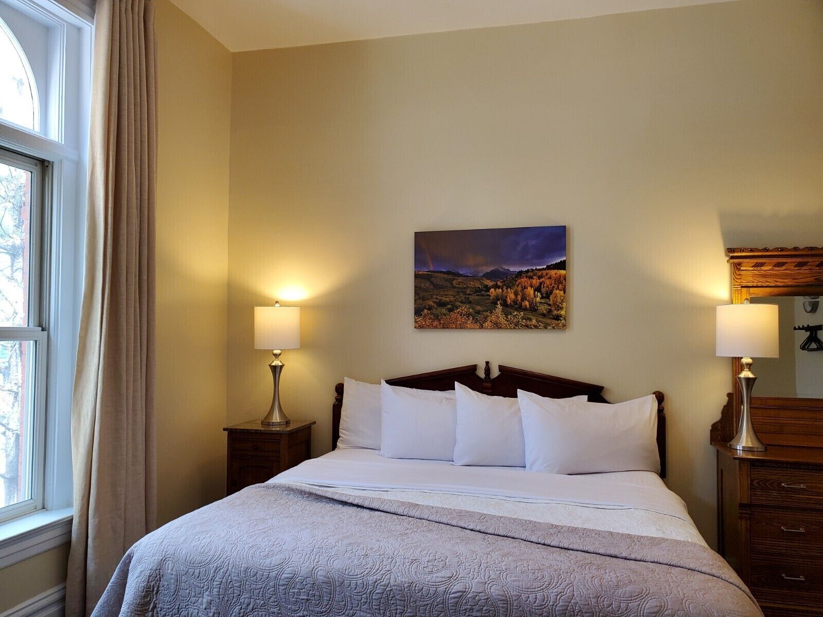A neatly made bed with white linens and a gray quilt, flanked by two bedside tables with lamps, a painting of a landscape on the wall, and a window with curtains.