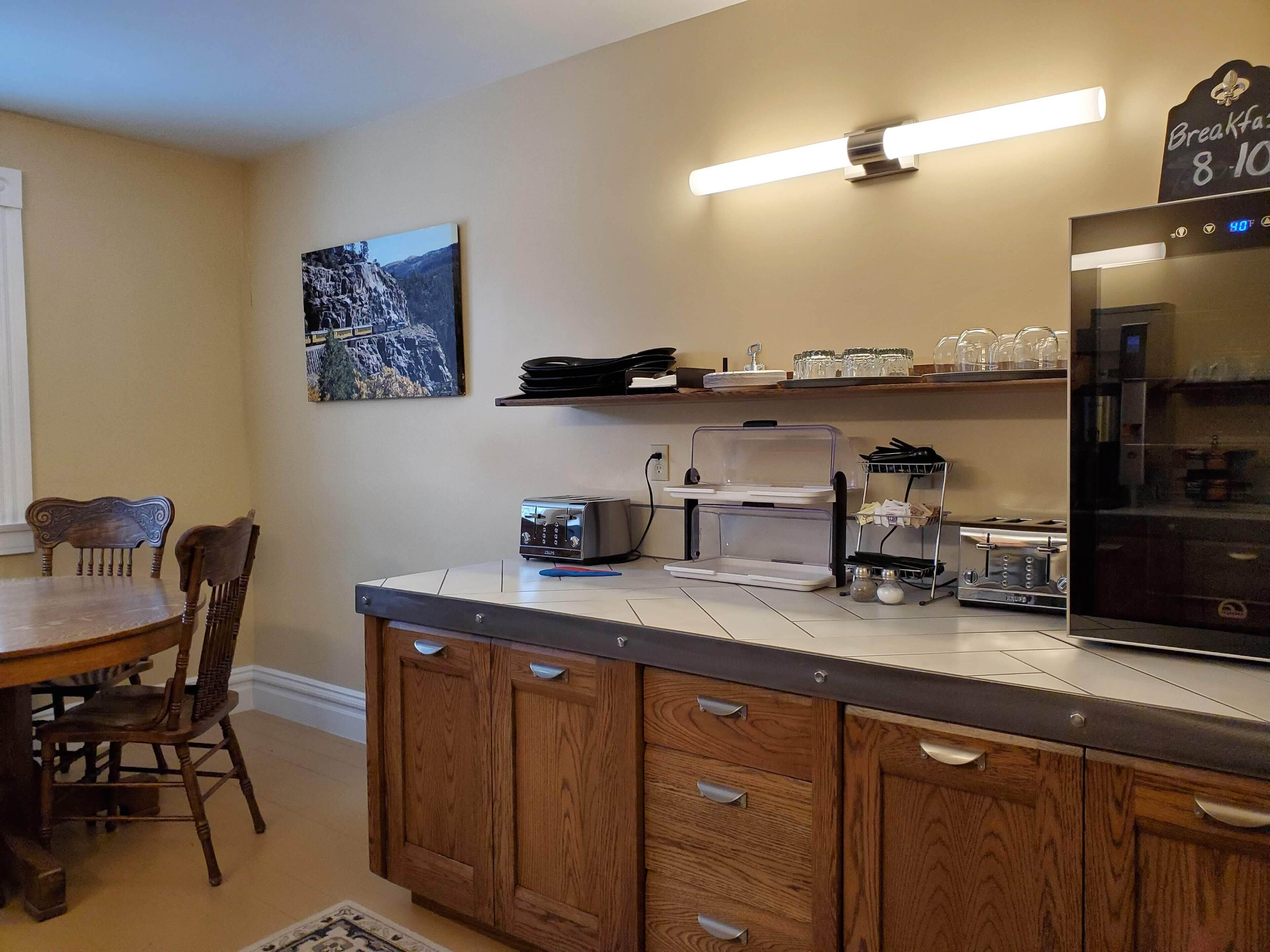 A kitchen with wooden cabinets, a countertop with a toaster, a coffee maker, and a refrigerator. There is a dining table with chairs and a wall-mounted shelf with dishes.