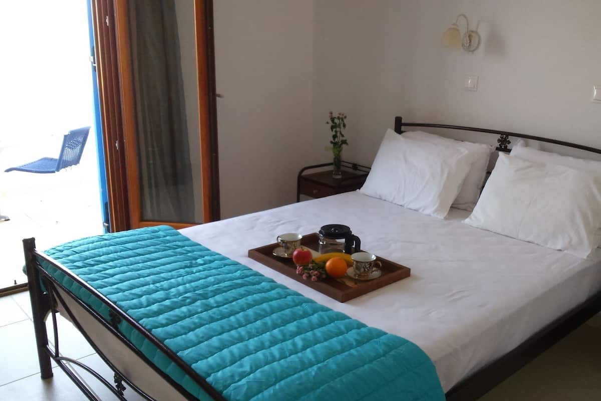 Un lit bien fait, recouvert d’une courtepointe turquoise, une table avec des fruits et une cafetière, et une table de chevet ornée d’un vase.