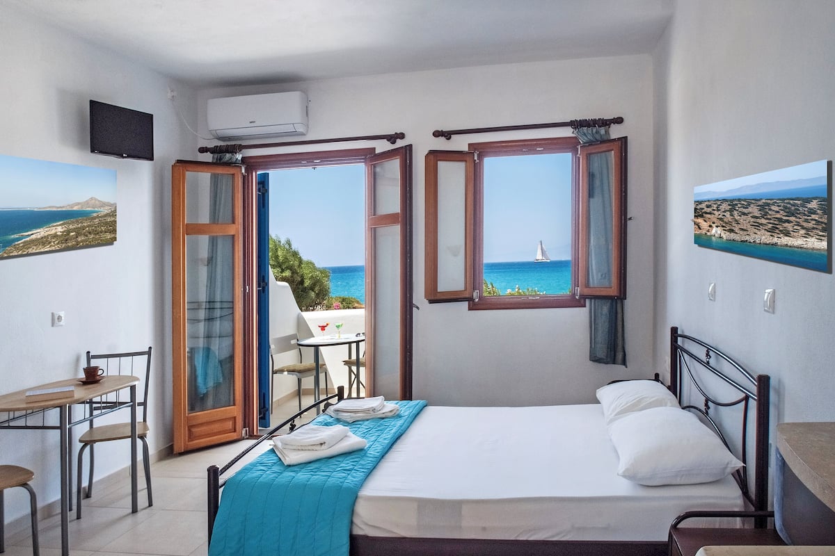 Une chambre à coucher avec un lit, un bureau et des chaises, une télévision, et une vue sur la mer par de grandes fenêtres ouvertes.