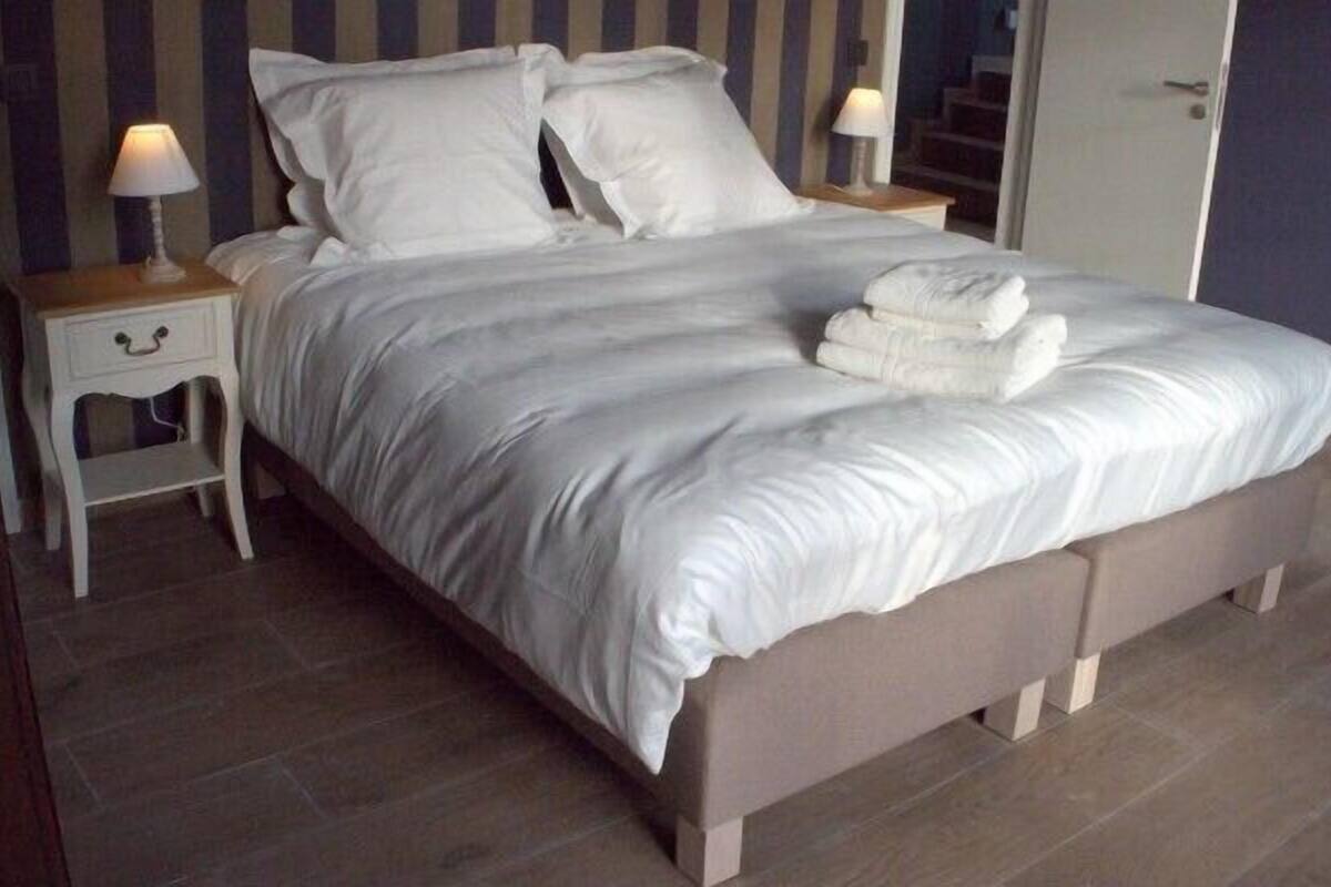 Un lit bien fait, avec du linge de lit blanc et une serviette pliée posée sur le côté. Il y a deux tables de chevet avec des lampes et un petit tiroir. La pièce a un plancher en bois et un mur à rayures au fond.