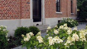 A brick building with a black door and windows, surrounded by a garden with white flowers. - Hotel Vorsen (Gingelom)