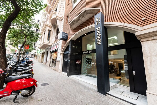 A hotel entrance with a modern facade, a clear sign indicating the name 'Rekord Hotel', and a row of parked scooters on the sidewalk.