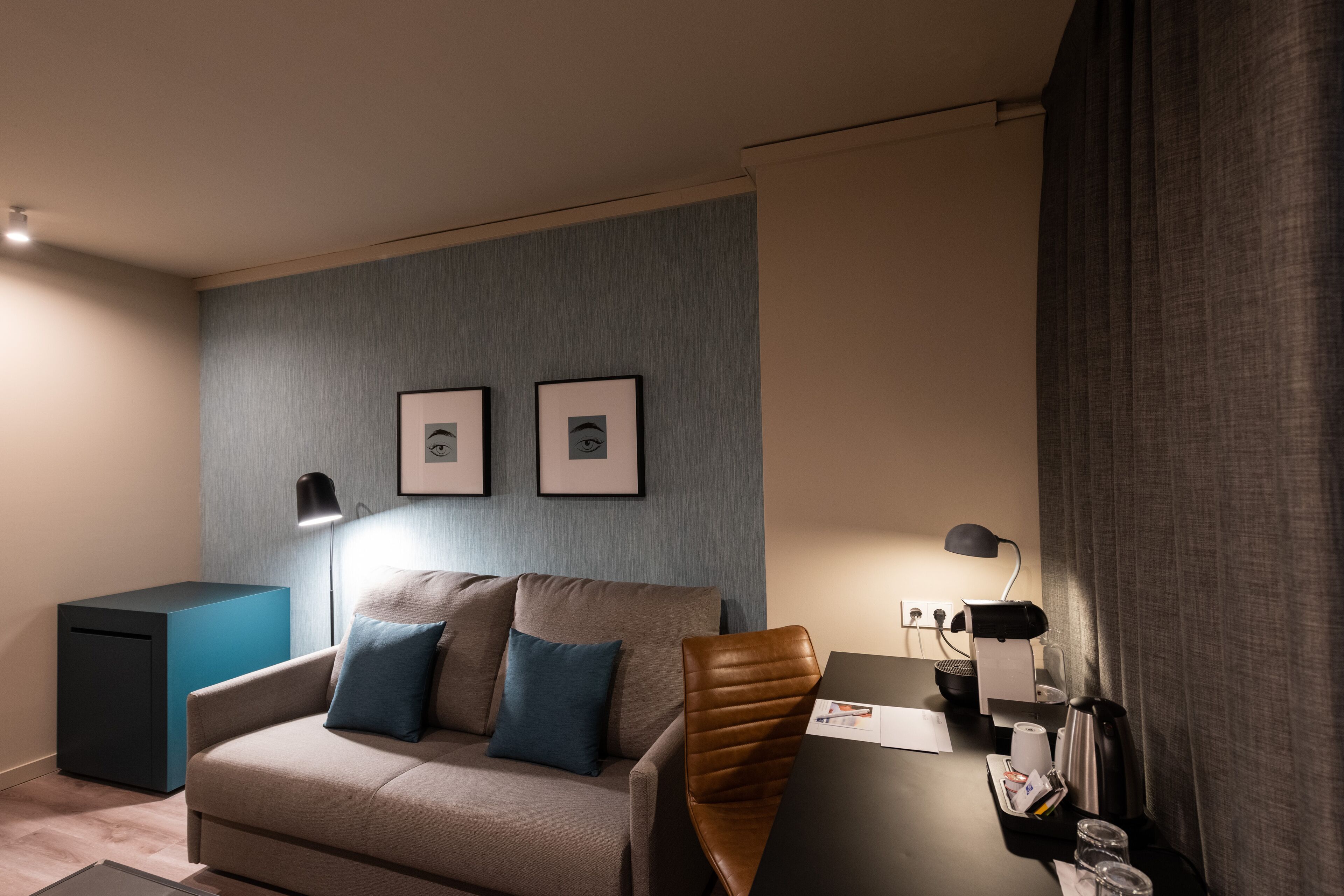 Une chambre d’hôtel moderne équipée d’un canapé, d’un bureau avec une chaise, d’une machine à café et de tableaux encadrés au mur.