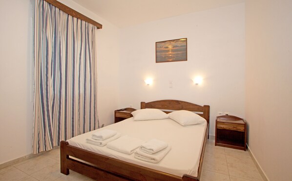 A neatly made bed with white linens and towels, a wooden headboard, two bedside tables with lamps, and a painting of a sunset on the wall. - Joanna's Apartments (Naxos)