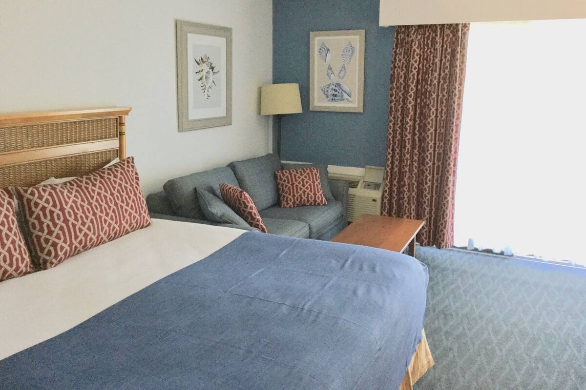 A hotel room with a large bed, a sofa, a small table, and a desk. There are two framed pictures on the wall.