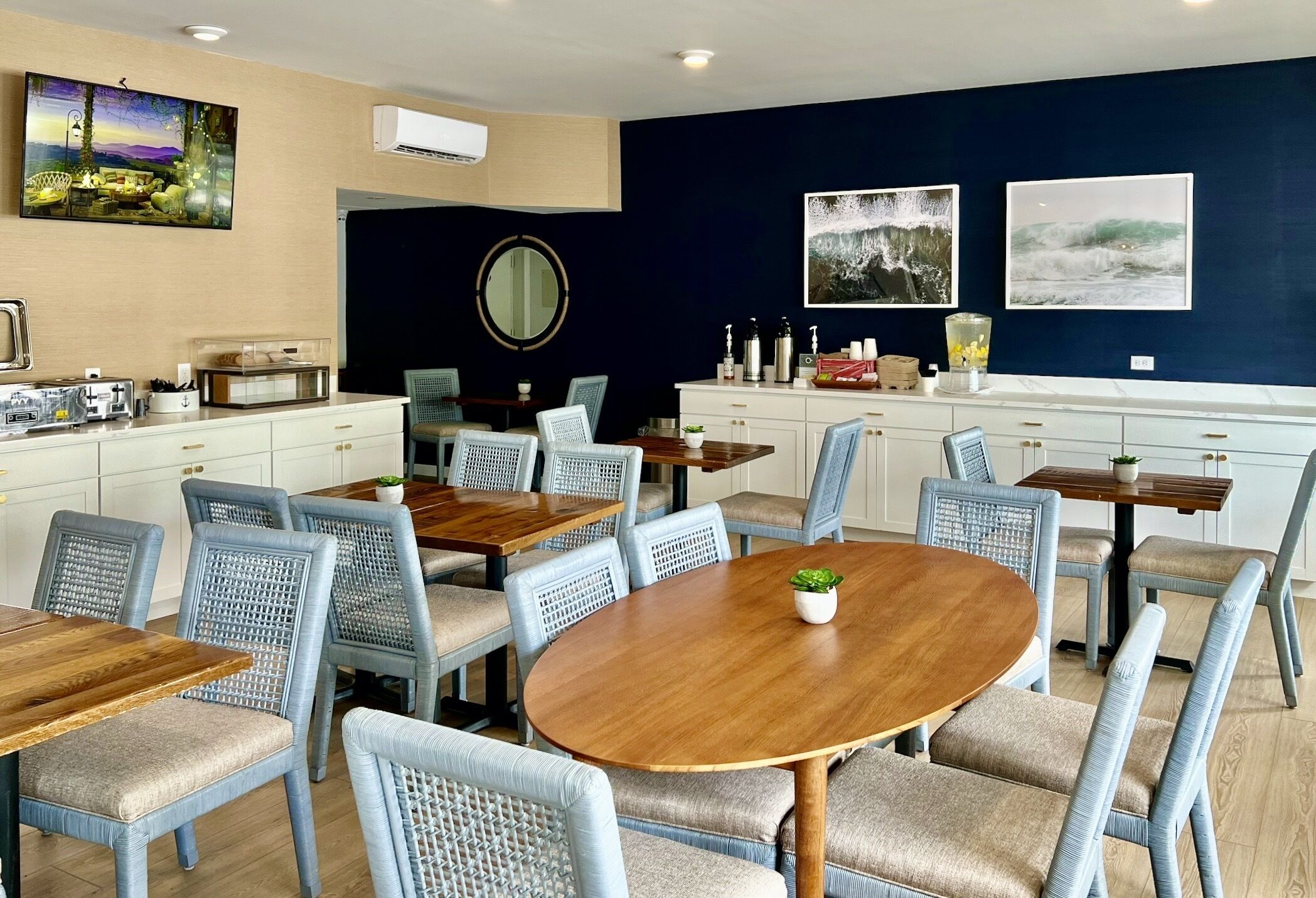 Un comedor con mesas redondas y sillas tapizadas en azul, un armario blanco con electrodomésticos de cocina, y obras de arte en las paredes.