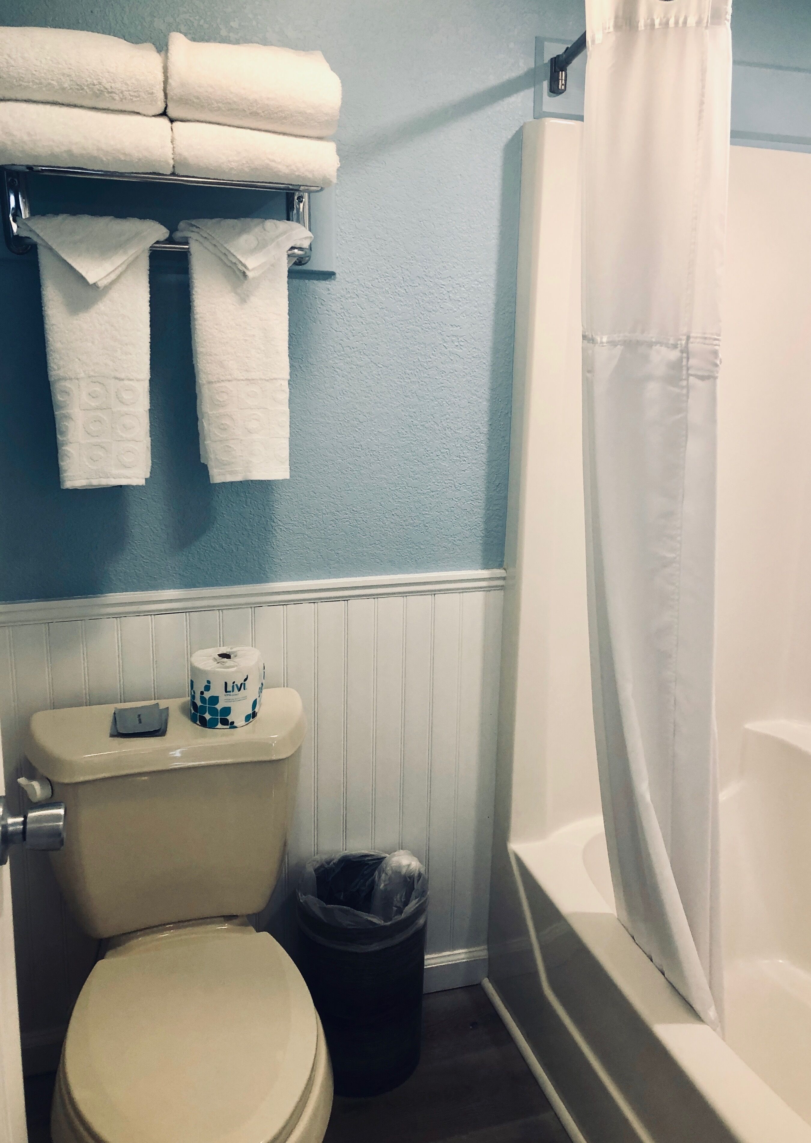 A bathroom with a toilet, a trash can, a shelf with folded towels, and a bathtub with a shower curtain.