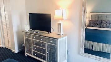 A hotel room with a TV on a wooden cabinet, a bedside lamp, a bed with patterned pillows, and a large mirror.