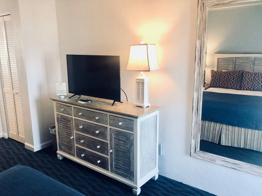 Ein Hotelzimmer mit Fernseher auf einer Holzkommode, einer Nachttischlampe, einem Bett mit gemusterten Kissen und einem großen Spiegel.