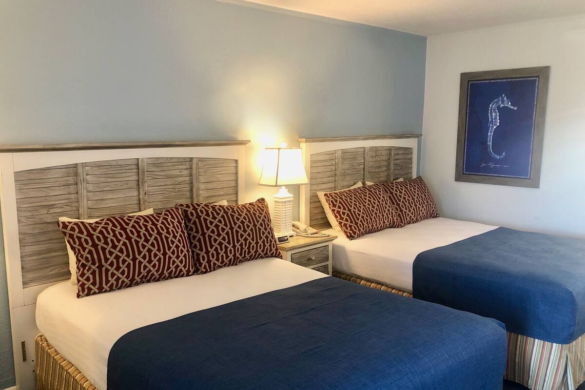 A hotel room with two beds, each with a wooden headboard, a nightstand with a lamp, and a framed seahorse artwork on the wall.