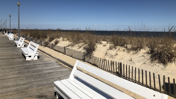Une promenade en bois avec des bancs blancs et une plage de sable bordée de dunes.