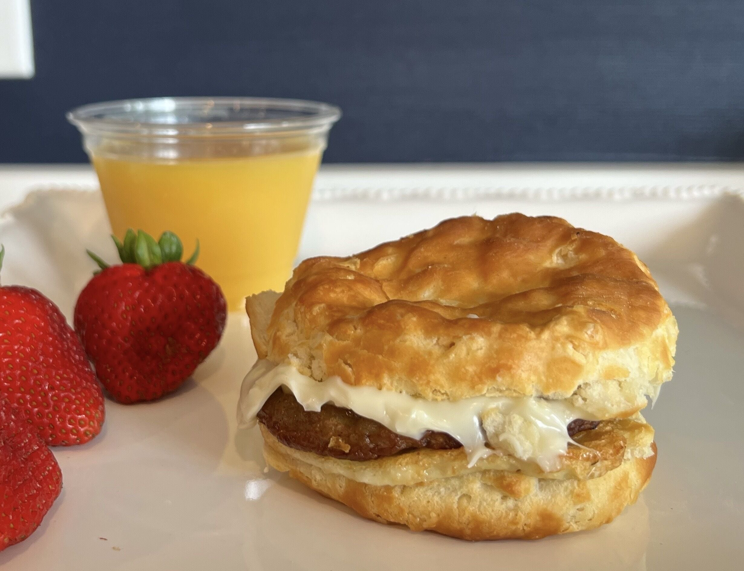 A breakfast sandwich on a white plate with two strawberries and a glass of orange juice.
