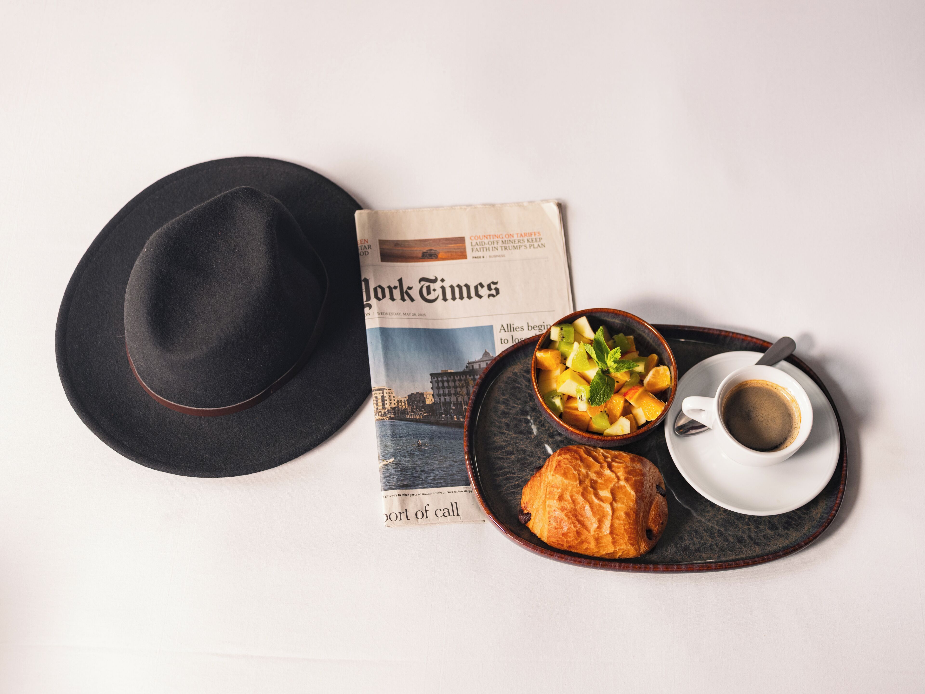A newspaper, a black hat, a bowl of fruit, a croissant, and a cup of coffee on a tray.