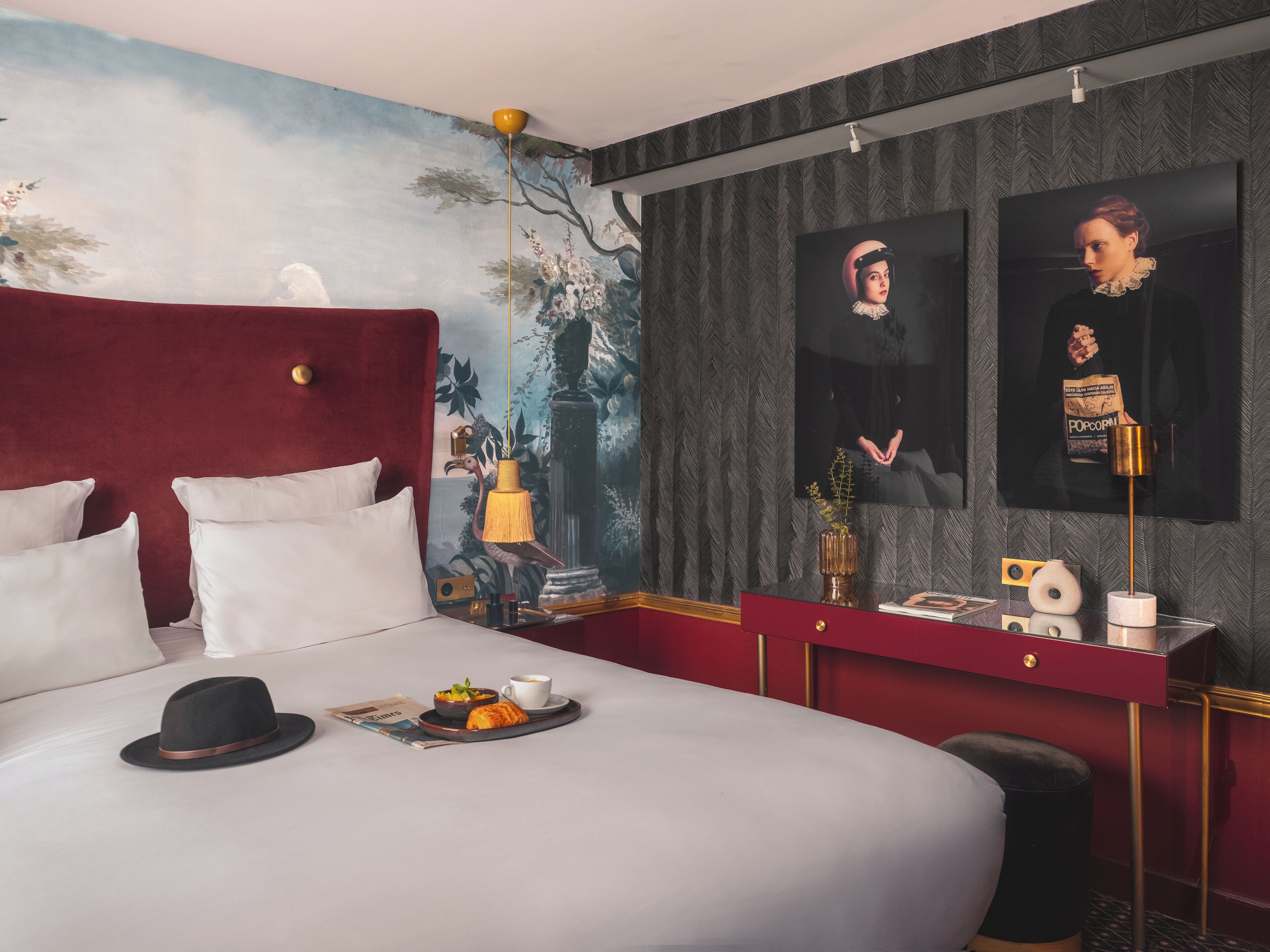Une chambre d’hôtel avec un grand lit, une tête de lit rouge, une fresque murale, un miroir et une petite table sur laquelle se trouvent un chapeau, un croissant et une tasse de café.