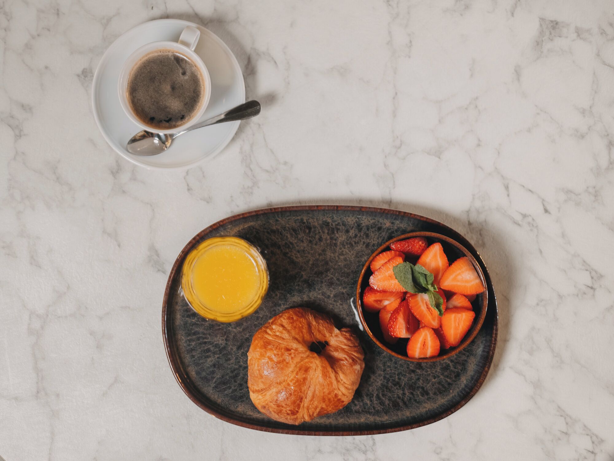 A breakfast tray with a croissant, a bowl of strawberries, and a cup of coffee on a marble surface.